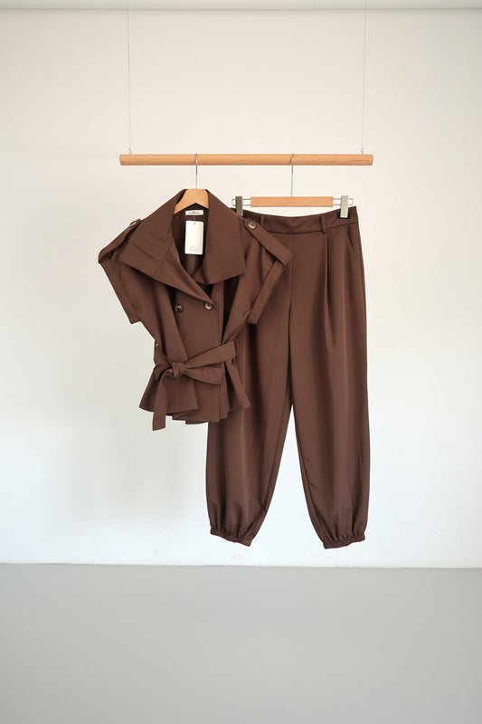 A brown two-piece clothing set consisting of a belted double-breasted wrap jacket and matching wide-leg trousers, hanging on black hangers against a white paneled door.