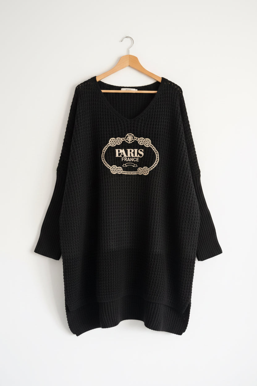 A black waffle-knit long-sleeve sweater with a beige 'PARIS FRANCE' decorative graphic on the chest, hanging against a white paneled wall.