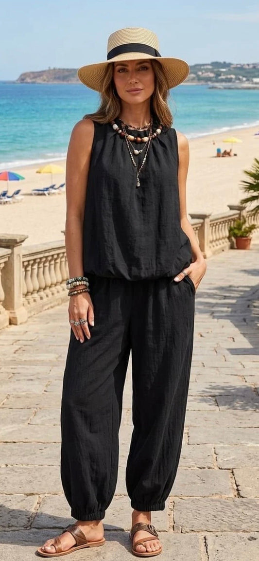 A woman stands on a stone walkway by the beach wearing a sleeveless black linen-like top and matching loose black pants, with a wide-brim beige hat, layered necklaces, bracelets, and brown sandals.