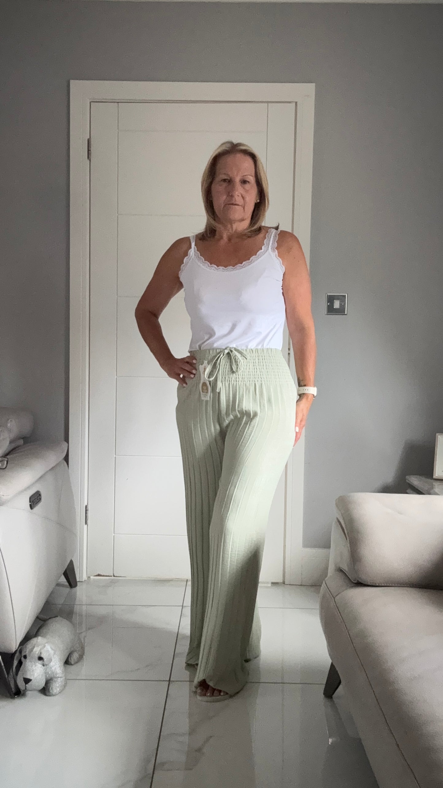 Woman in a white top and light green pants standing in a room with a white door and gray walls.