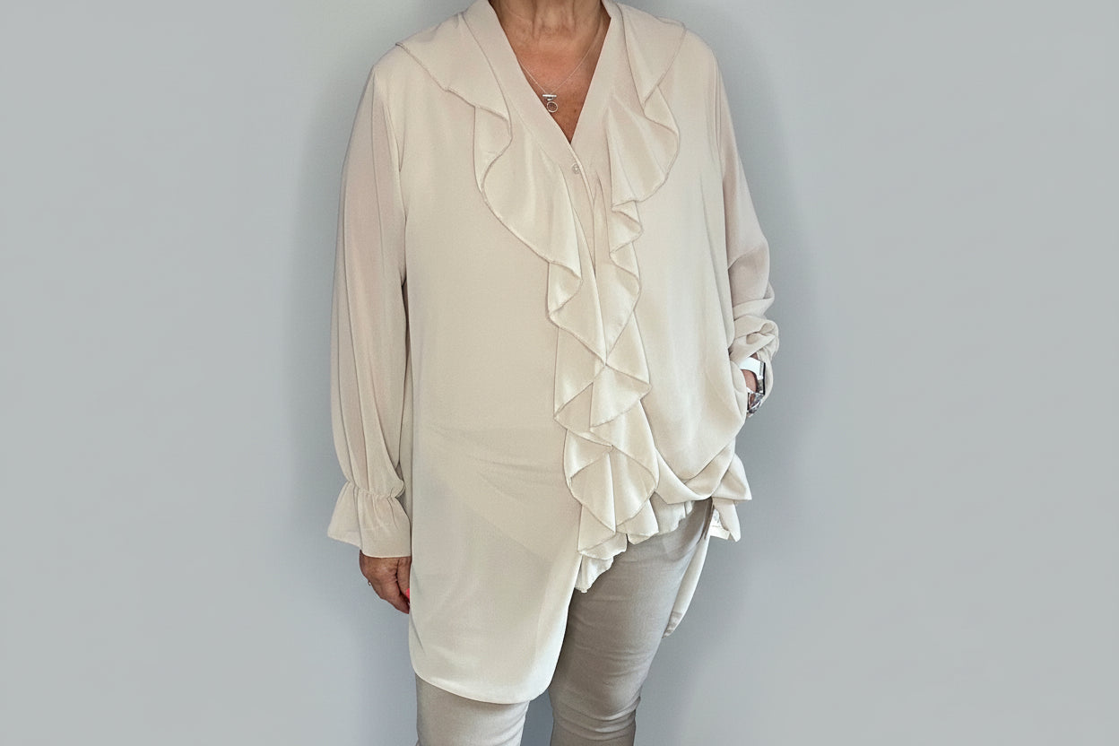 Person wearing a beige blouse with ruffles and light gray pants against a plain background