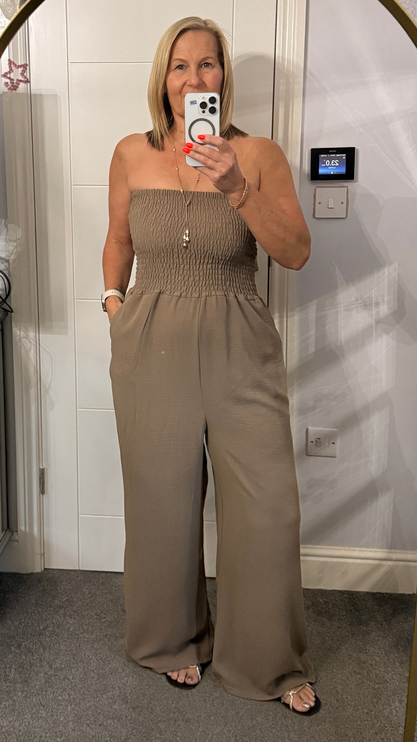 Woman taking a mirror selfie wearing a strapless jumpsuit in a room with white walls.