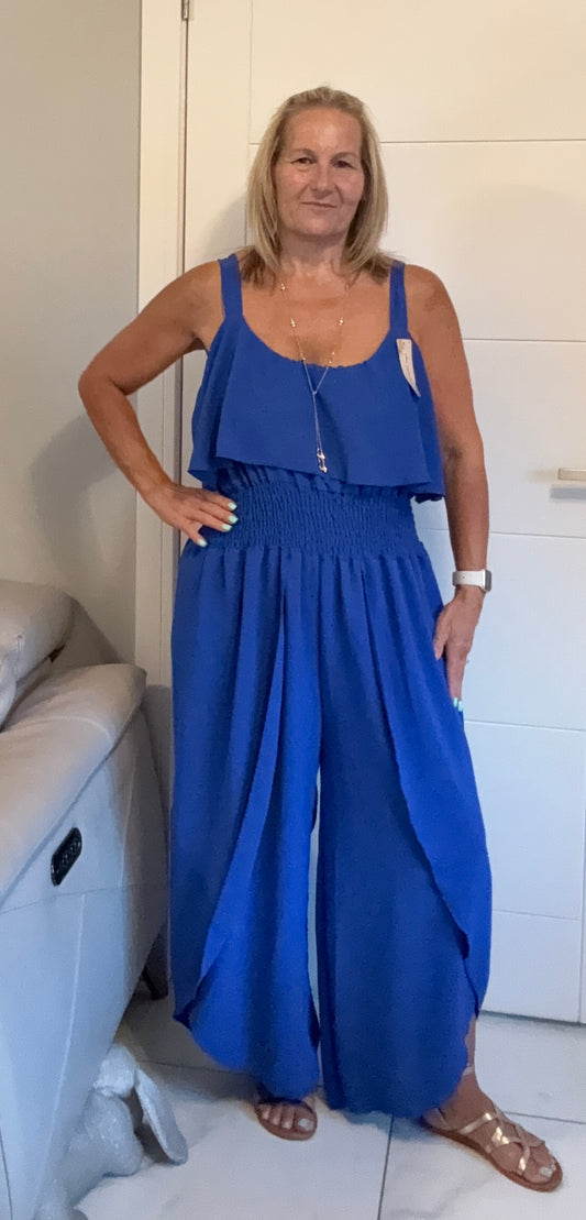 A woman modeling a royal blue sleeveless jumpsuit with a smocked waist and wide legs, standing in a room with white paneled walls.