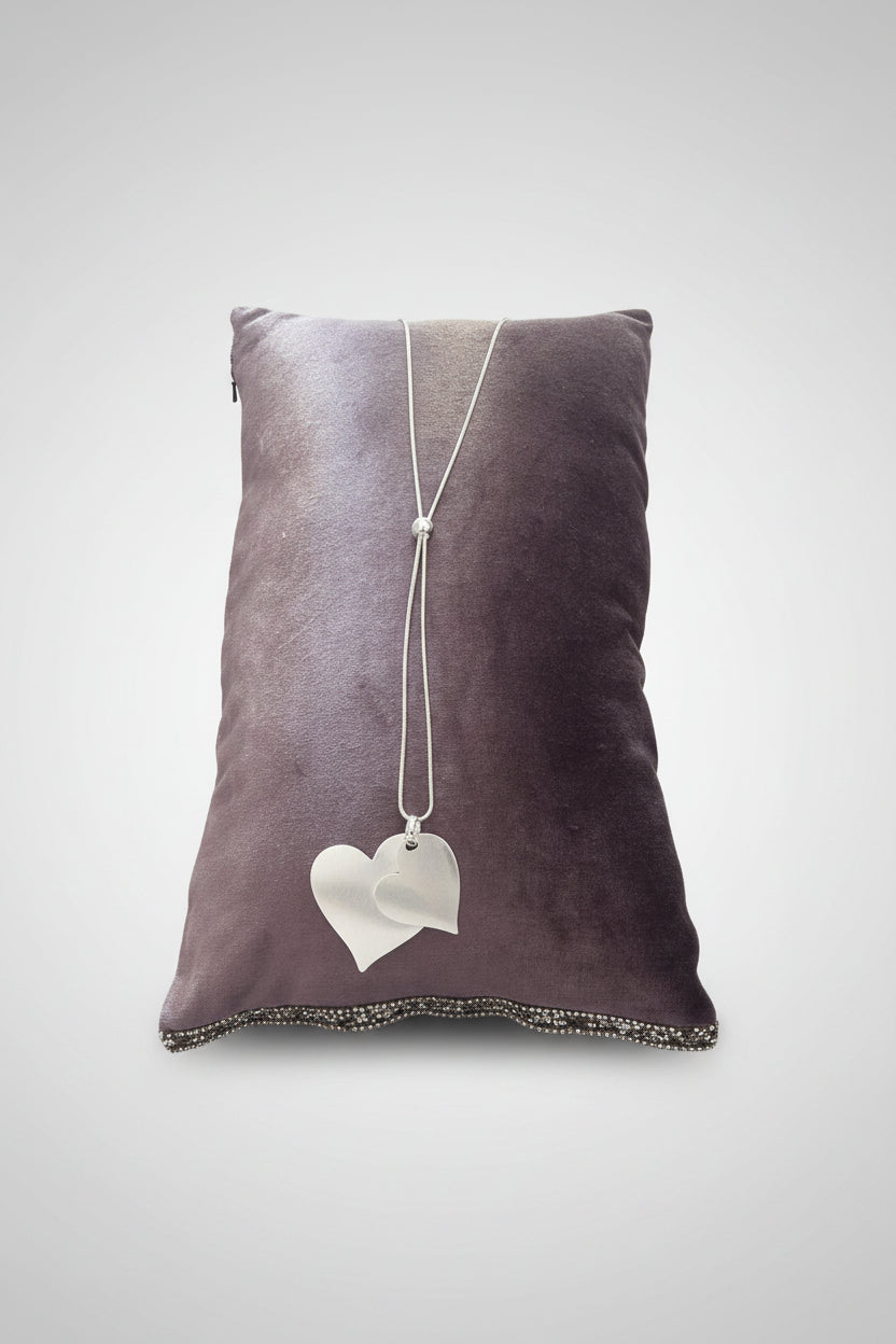 A silver-toned long necklace with two interlocking heart-shaped pendants, displayed on a dark brown velvet pillow over a gray stone surface.
