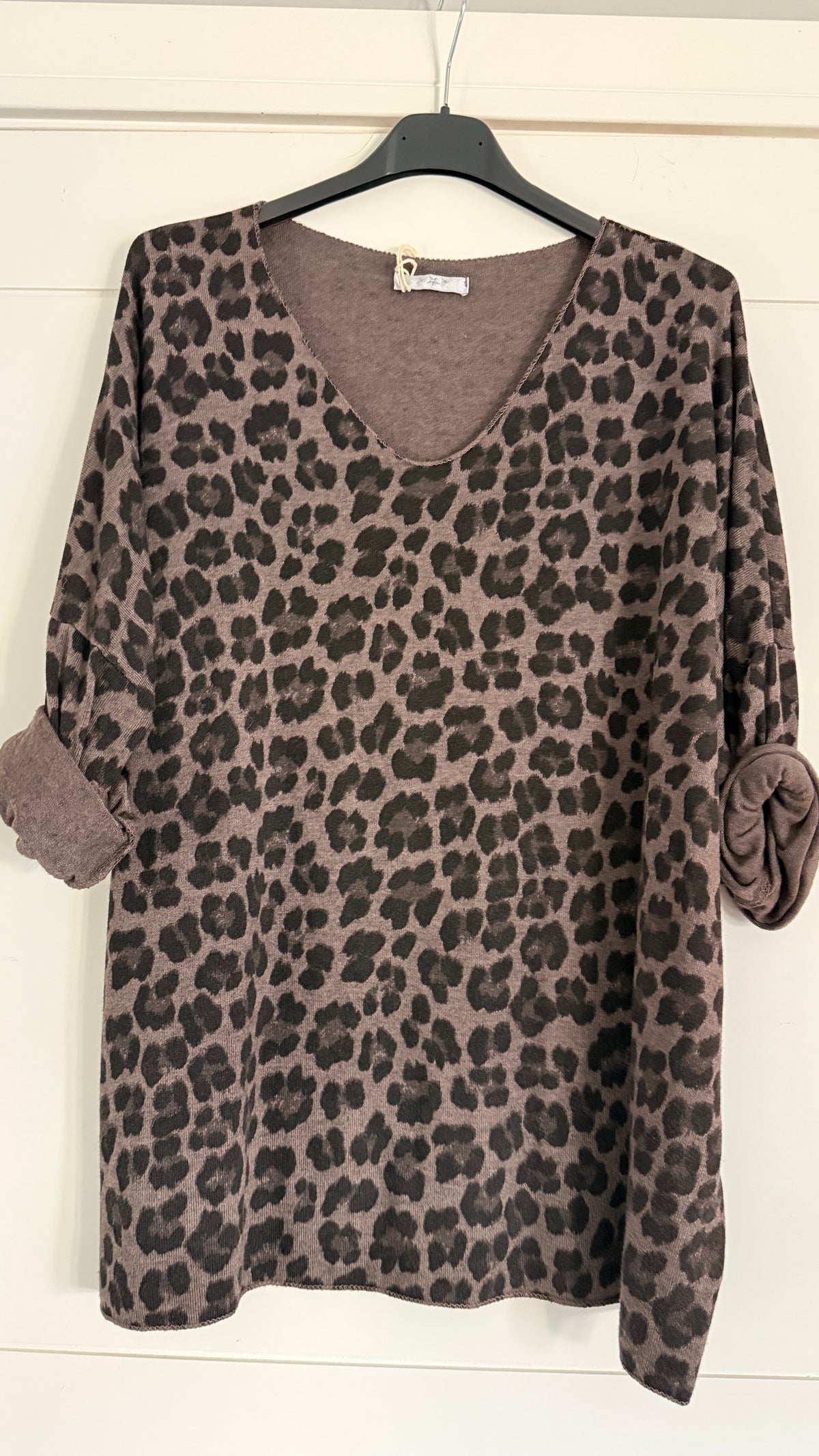 A leopard print long-sleeve top with rolled cuffs, hanging on a black hanger against a white paneled wall.