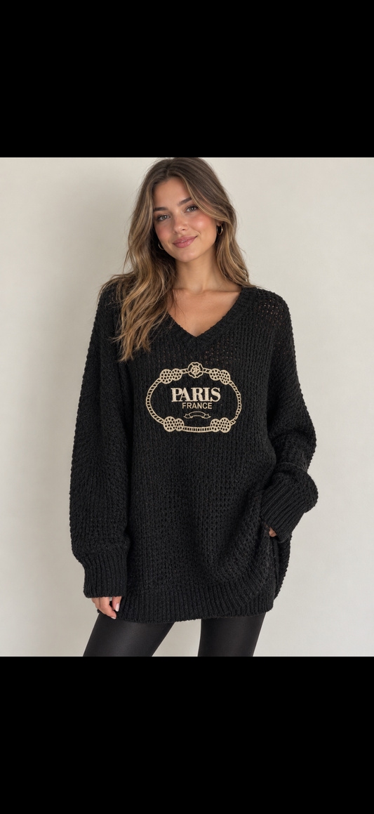 A black waffle-knit long-sleeve a women wearing sweater with a beige 'PARIS FRANCE' decorative graphic on the chest, hanging against a white paneled wall.