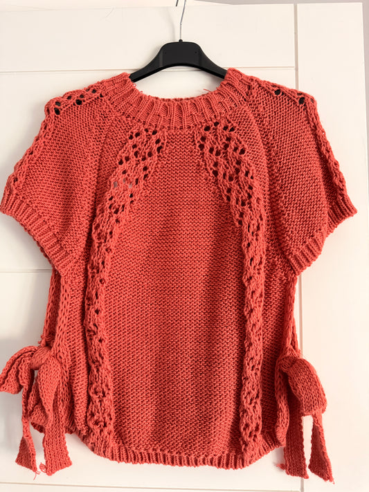 hand-knit short-sleeve top with a a coral textured lace panels and side ties, hanging on a black hanger against a white paneled background.