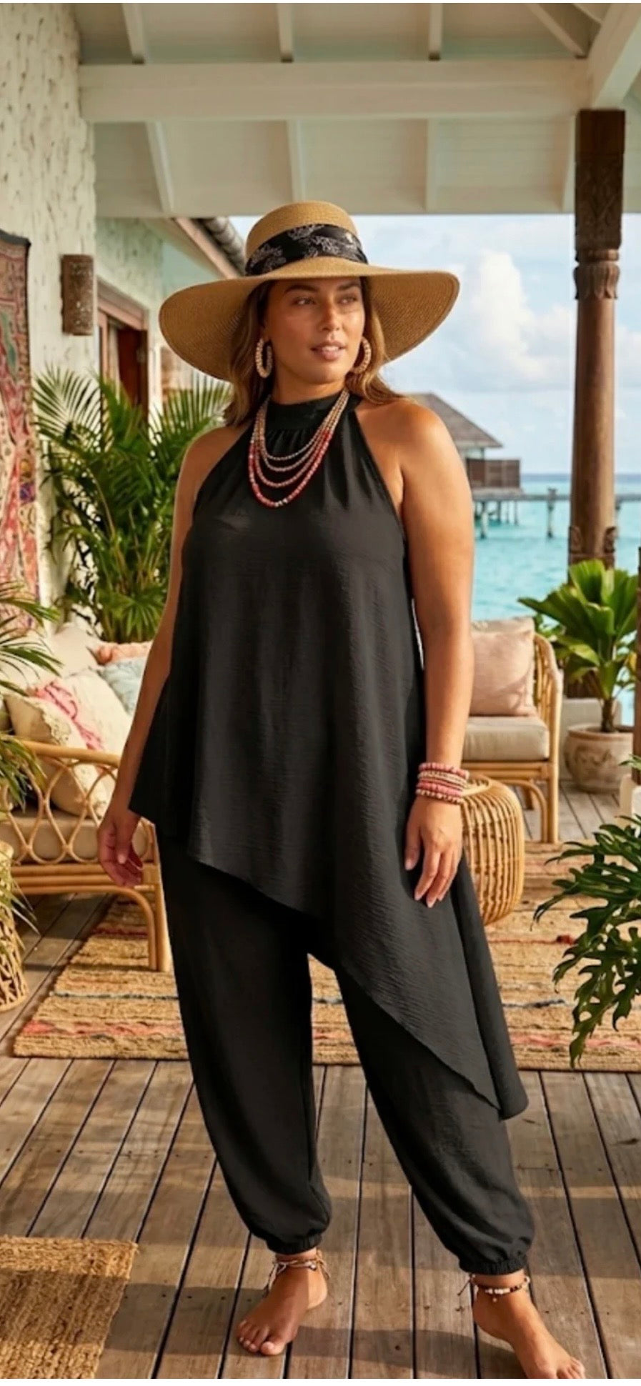Woman in a black outfit with a wide-brimmed hat standing on a wooden deck by the ocean.