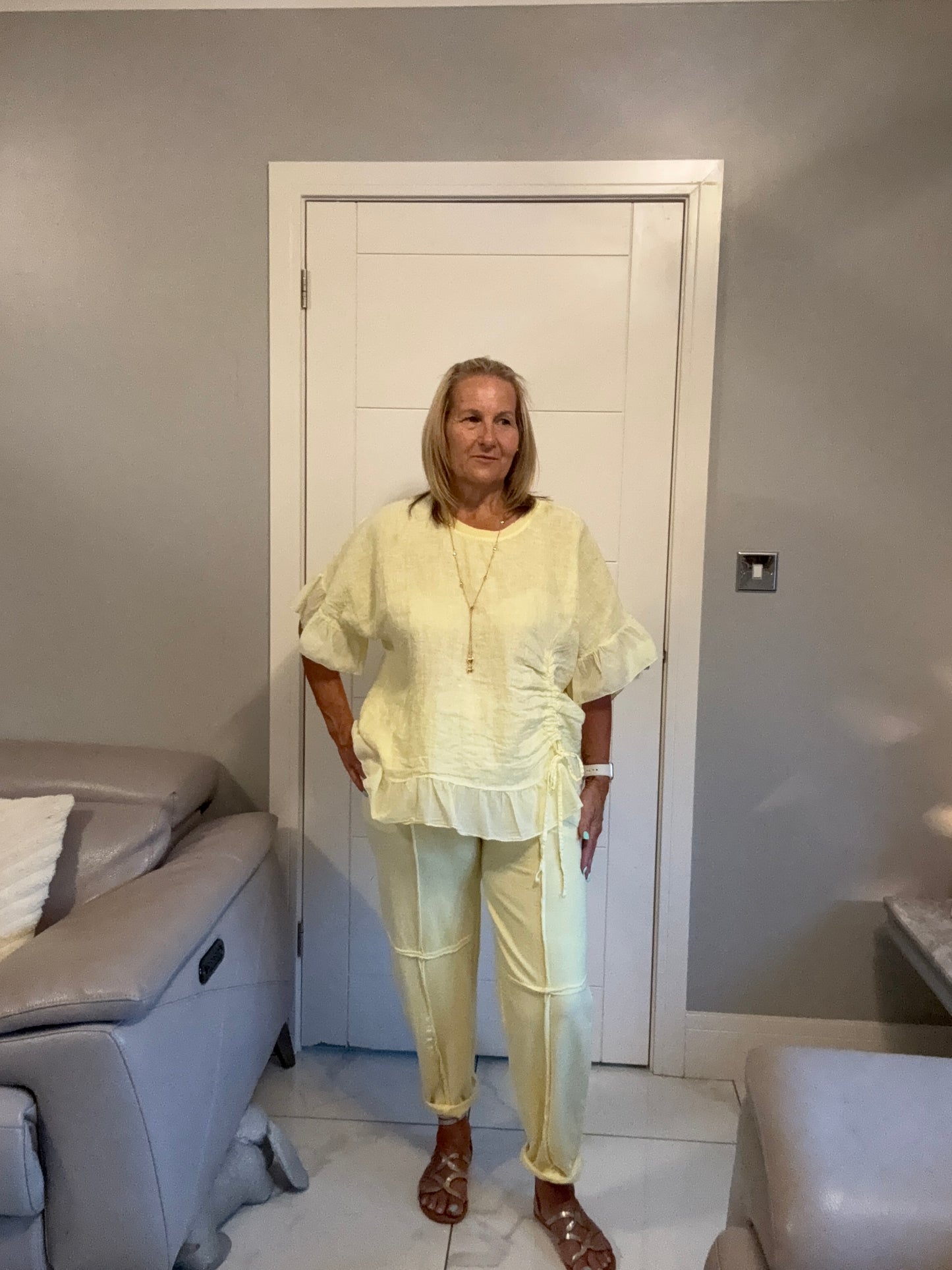 A woman wearing a light yellow matching two-piece set consisting of a short-sleeve ruffled blouse and trousers, standing in a modern room with a white door and gray walls.