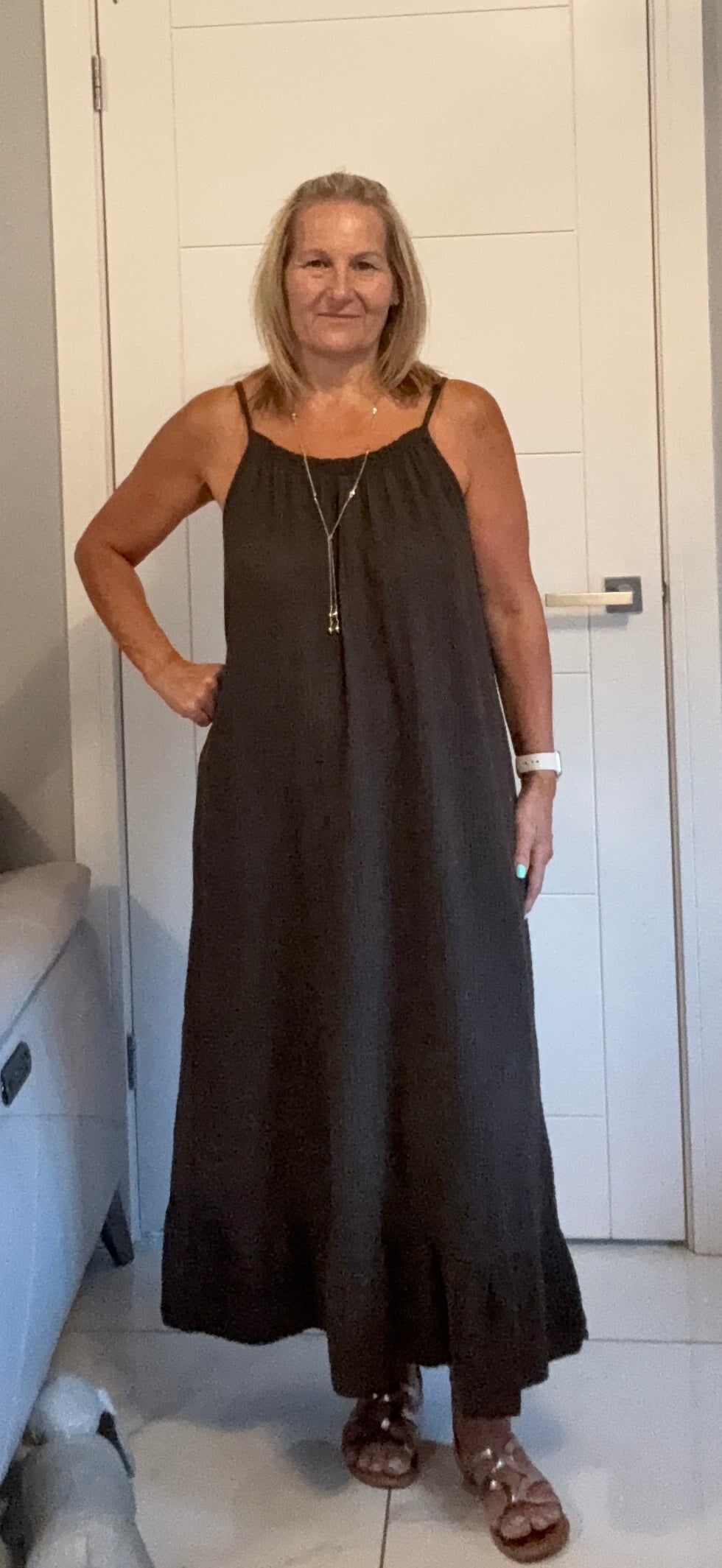 A woman modeling a dark brown sleeveless maxi dress with thin straps and a long necklace, standing in a home setting with a white door and tiled floor.