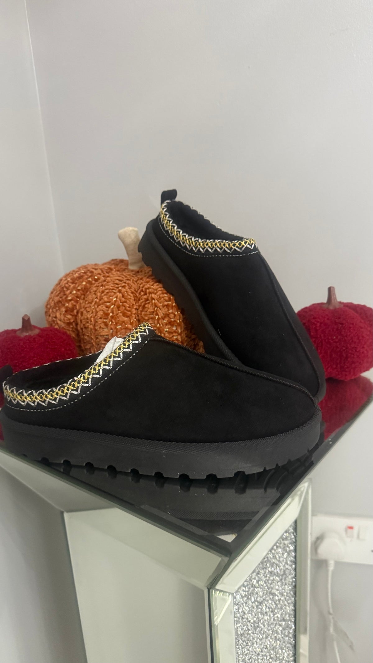 Black slipper with decorative trim on a reflective surface with pumpkins in the background