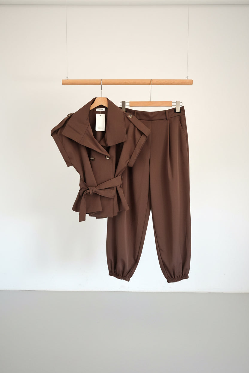 A two-piece women's outfit in a solid dark brown color, hanging on a wooden hanger bar. The set includes a belted wrap-style short-sleeve top and matching high-waisted cropped pants with elastic cuffs, set against a plain white wall.