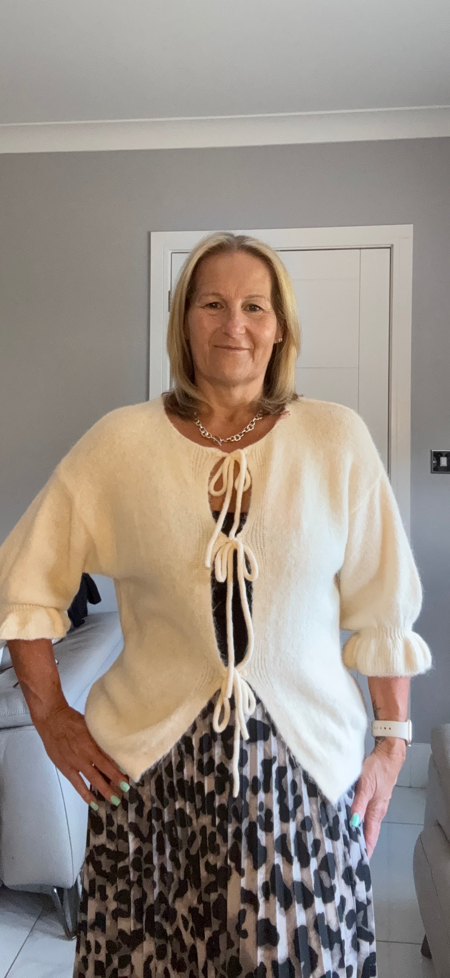 A woman wearing a cream-colored knit cardigan with tie-front details and a black-and-white leopard print skirt, standing in a home interior setting.