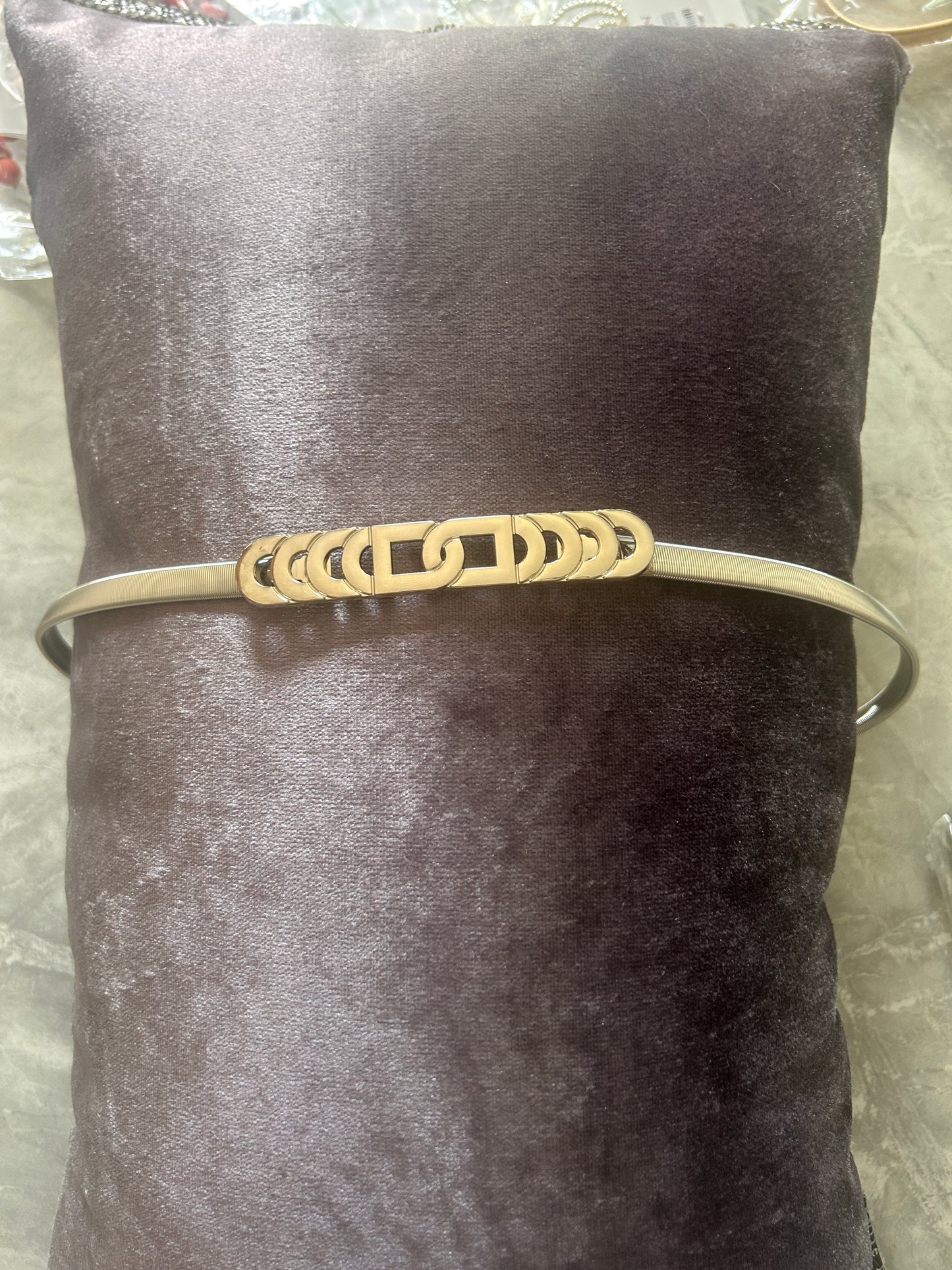 A silver-toned metal bel with a decorative interlocking link design, displayed on a dark brown velvet pillow.
