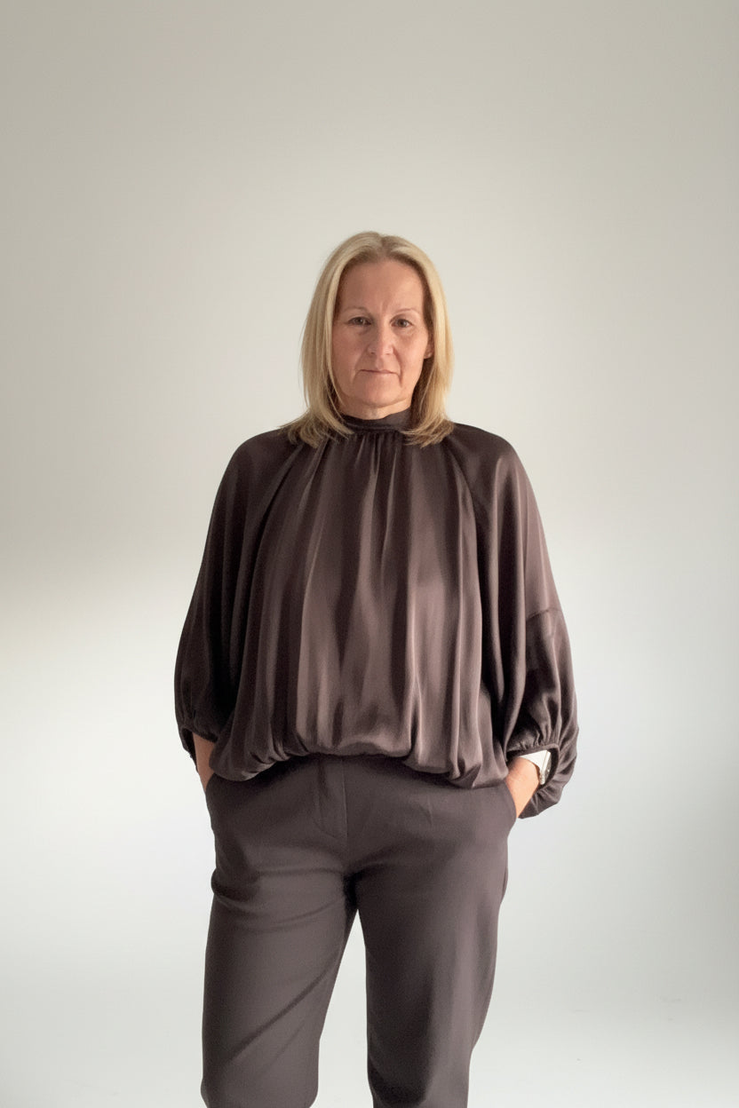 A woman wearing a dark brown satin-like blouse with voluminous balloon sleeves and matching dark brown trousers, standing in a living room setting.
