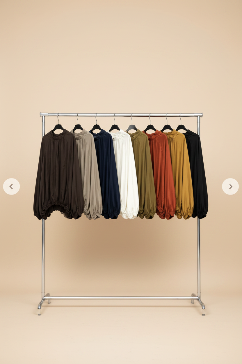A metal clothing rack displaying a row of loose, long-sleeve bubble-sleeve tops in multiple colors: brown, taupe, navy, white, olive, rust, mustard, and black, set against a solid beige background.
