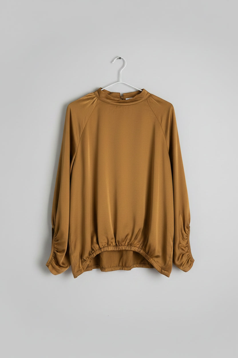 A loose, long-sleeve blouse in a warm camel brown or ochre color, hanging on a black hanger against a white paneled wall.