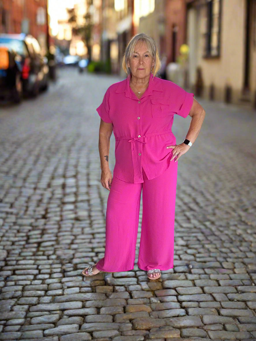 Woman wearing a bright pink outfit standing in a room with a mirror and various items on the floor.