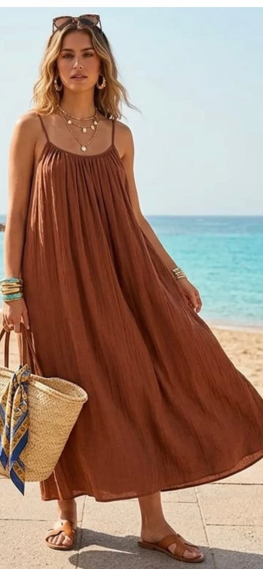 A woman wearing a flowing rust-brown sleeveless maxi dress stands on a beach with the ocean in the background. She is holding a woven straw tote bag and wearing layered necklaces and bracelets.