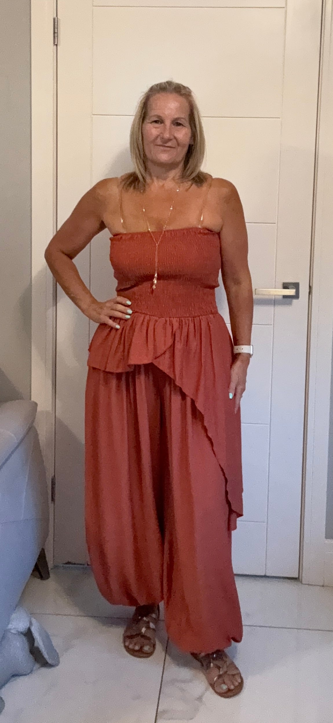A woman modeling a terracotta-colored strapless ruffled maxi dress with wide-leg harem-style pants, standing in front of a white door.