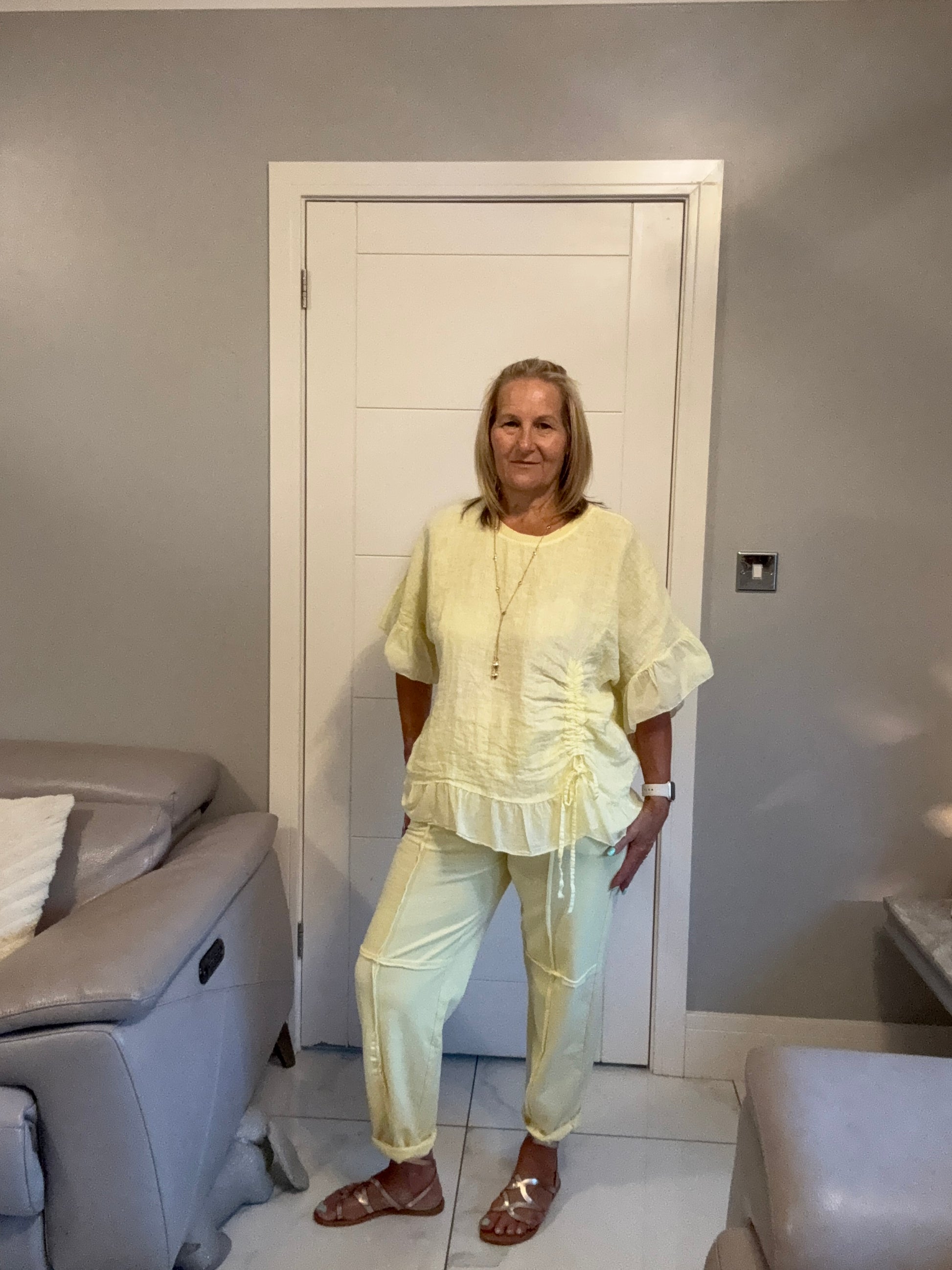 A woman wearing a pale yellow two-piece matching set with a ruffled long-sleeve top and straight-leg pants, standing in a room with a gray wall and white door.