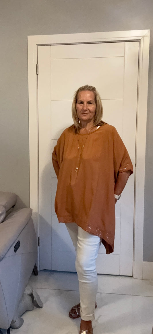 A woman modeling a loose-fitting, rust-brown short-sleeve tunic top with white pants, standing in front of a white door in an indoor room.