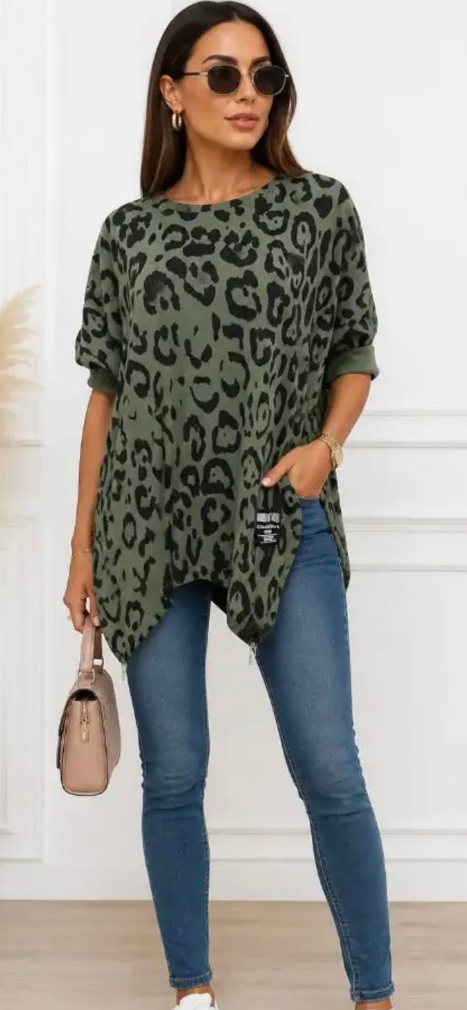 A woman modeling a short-sleeve olive green leopard print top with blue skinny jeans, sunglasses, and a beige handbag, standing against a white paneled wall.