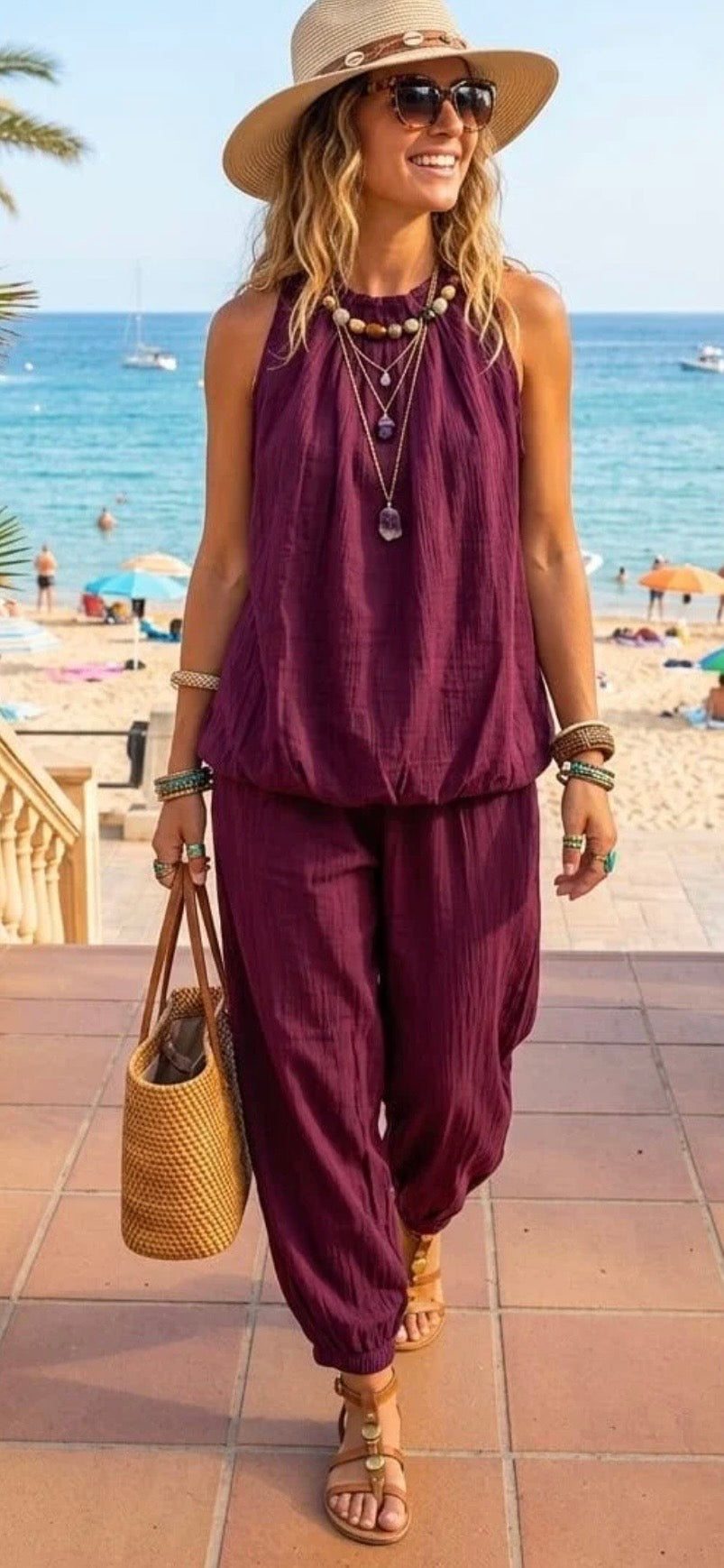 A woman walking on a beach boardwalk wearing a sleeveless deep purple harem-style two-piece set, a beige wide-brim hat, sunglasses, layered necklaces, and carrying a woven straw bag.
