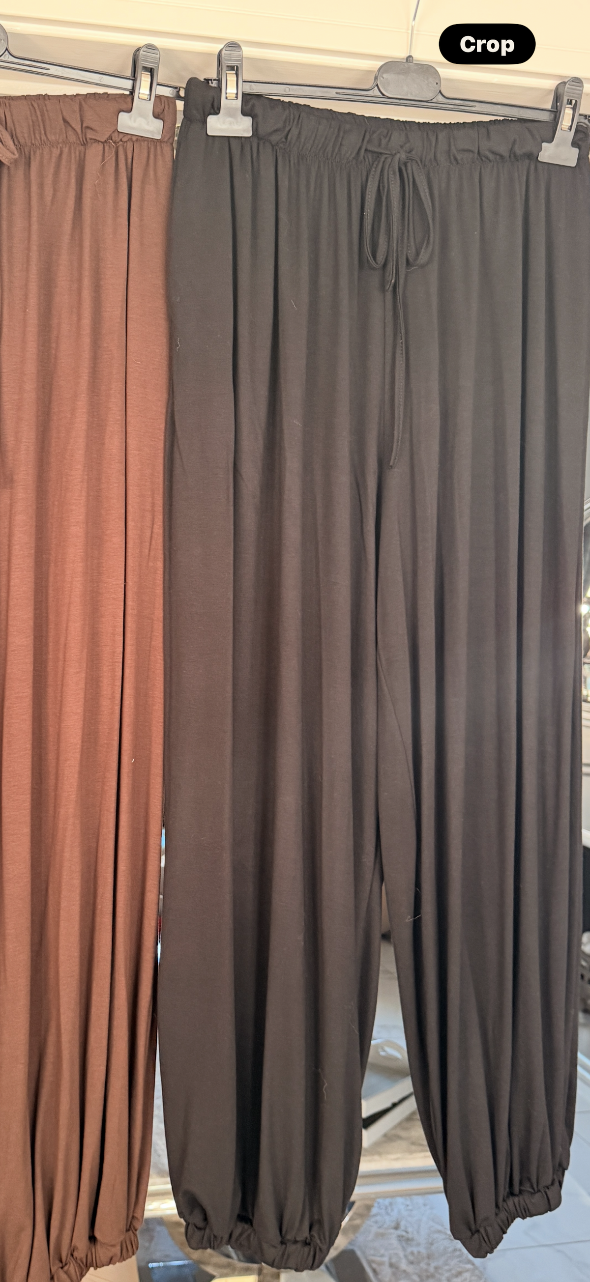 A pair of loose, drawstring-hem pants hanging on a hanger, shown alongside another pair in a different rust-brown color. The main pair is a dark charcoal gray.