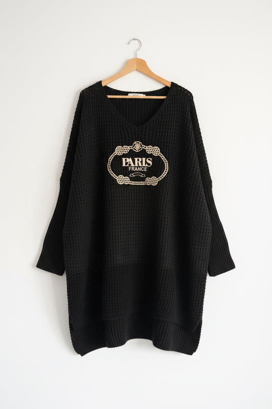 A black waffle-knit long-sleeve sweater with a beige 'PARIS FRANCE' decorative graphic on the chest, hanging against a white paneled wall.