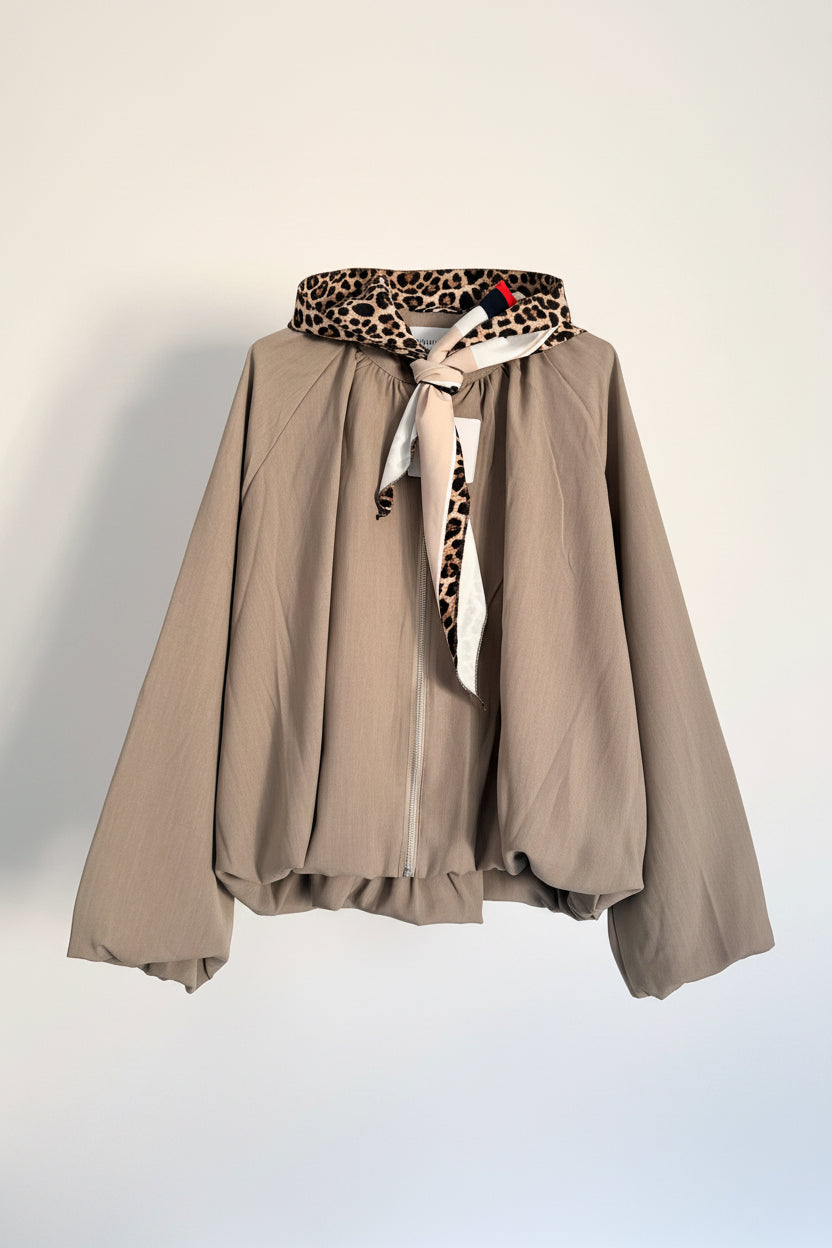 A taupe zip-up hooded jacket with a beige, black, and white patterned scarf tied around the collar, hanging against a white paneled wall.
