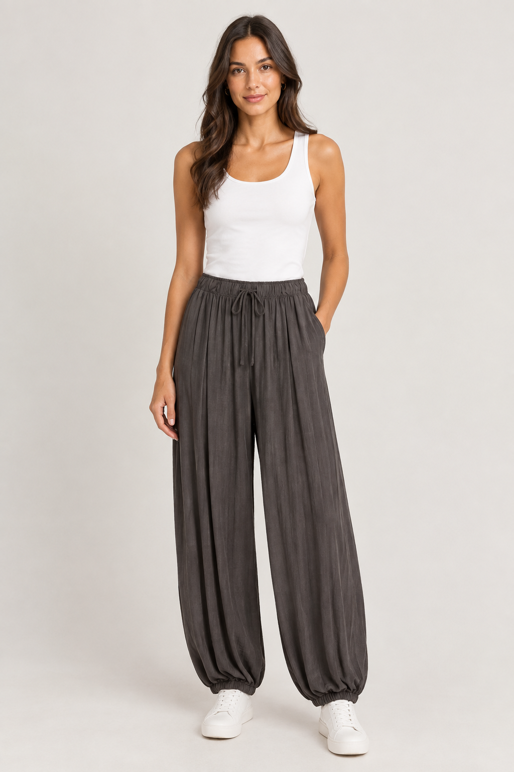 A woman modeling loose, high-waisted dark gray drawstring pants with a white tank top and white sneakers, standing against a light gray studio background.