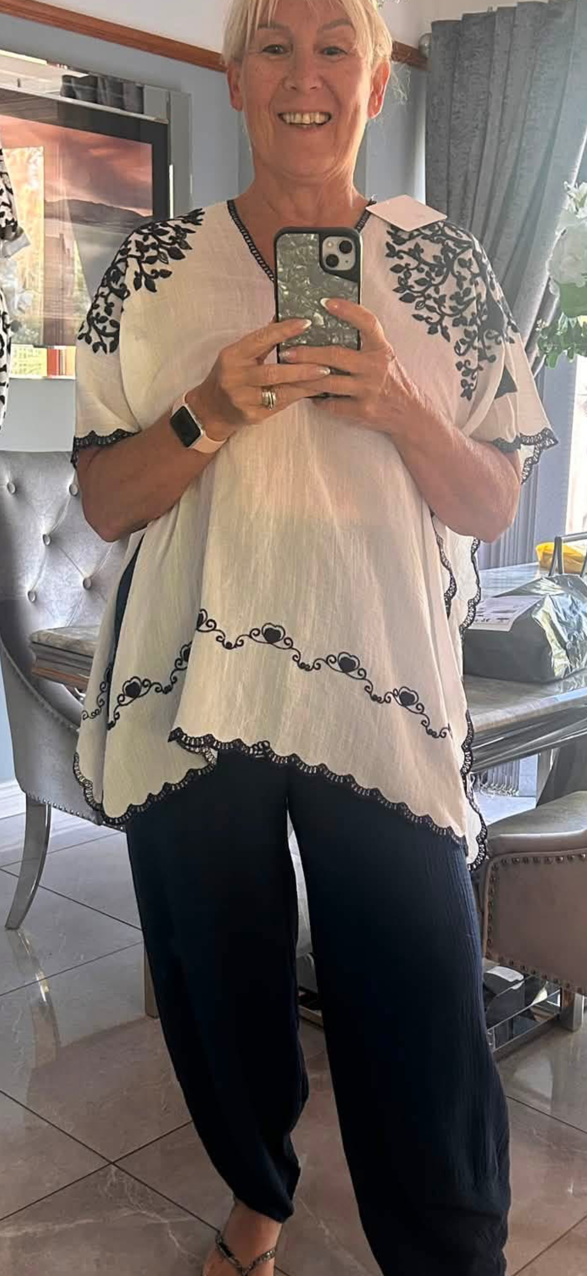 A woman taking a mirror selfie in a home setting, wearing a cream-colored short-sleeve blouse with black floral embroidery on the shoulders and hem, paired with dark navy pants.