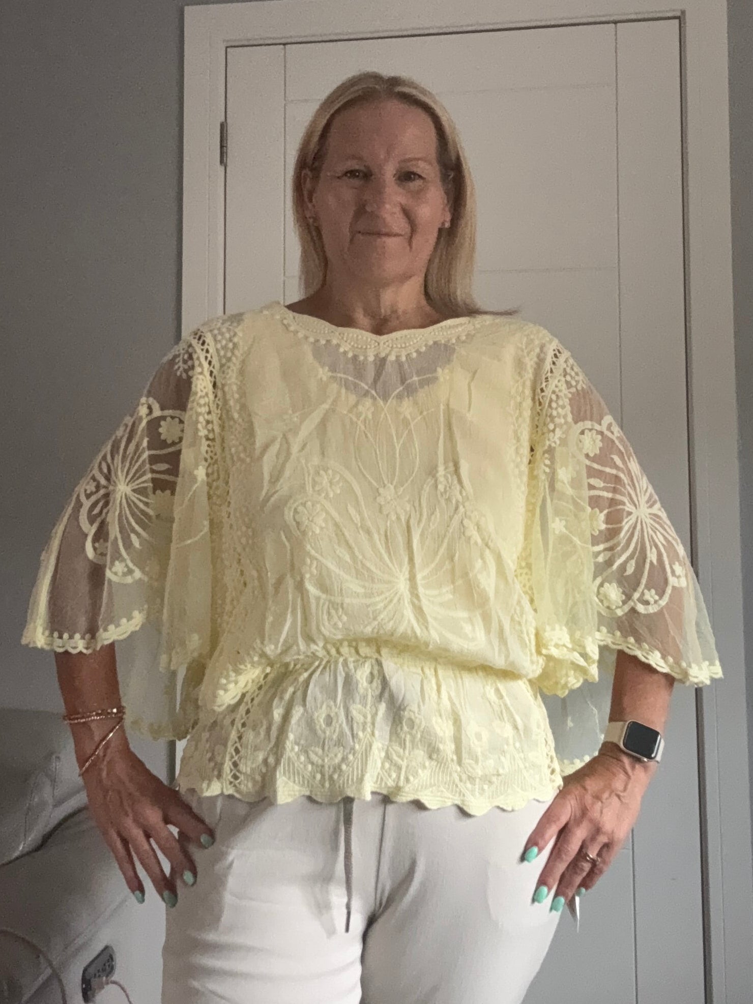 A woman wearing a pale yellow lace blouse with sheer flutter sleeves and white pants, standing indoors in front of a white door.