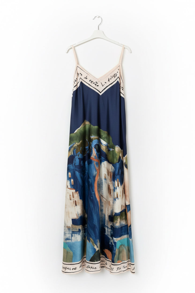 A sleeveless maxi dress with thin straps, featuring a navy blue upper section with cream lettering and a scenic landscape print in blue, green, and beige tones, hanging on a white hanger against a white paneled door.