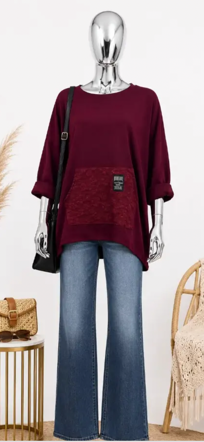 A mannequin styled in a burgundy knit top with a textured front pocket, paired with blue jeans and a black shoulder bag, set against a light studio background with decorative props.