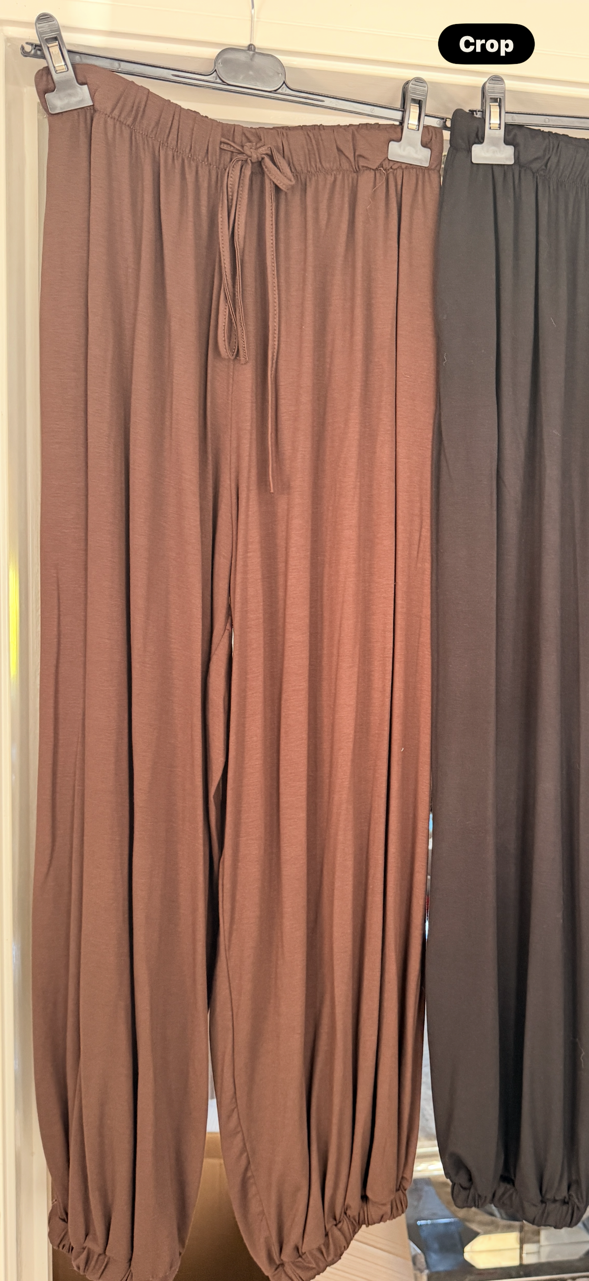 A pair of loose, elastic-waist jogger pants in a dusty rose-brown color hanging from a metal hanger, with a second pair in dark gray hanging beside it. The image has a 'Crop' label overlay in the top right corner.