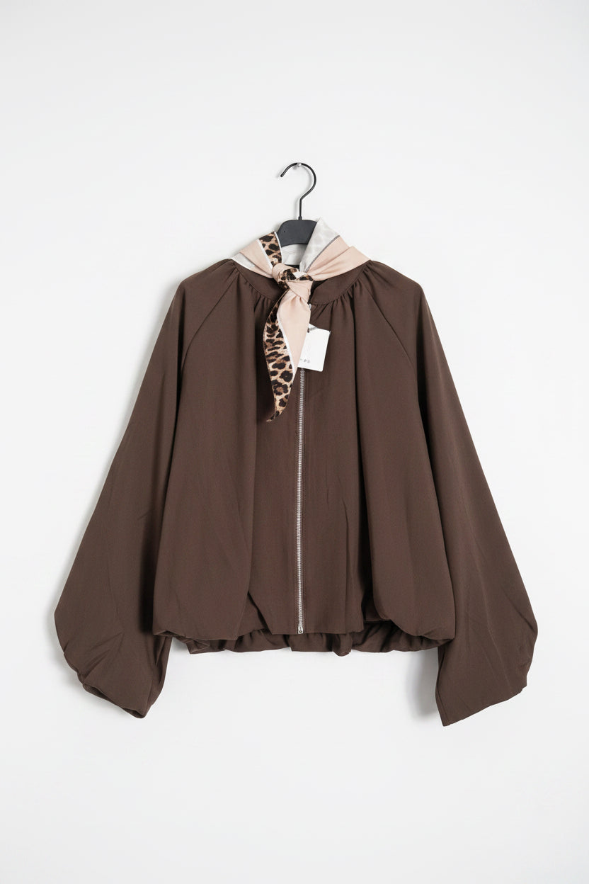 A brown long-sleeve zip-up jacket with a beige and leopard-print neck scarf, hanging on a white paneled wall.