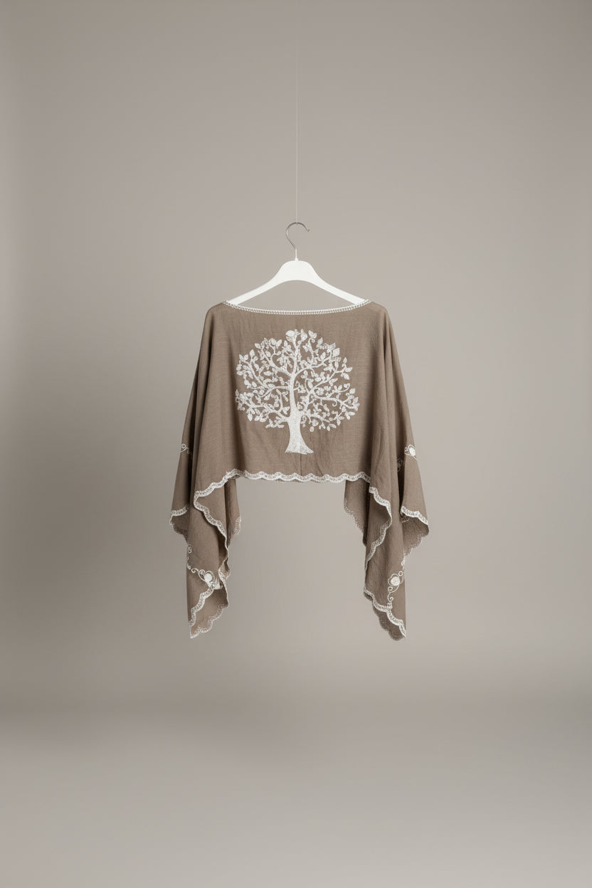 A taupe-brown draped poncho-style top with a large white embroidered tree of life on the front, hanging on a white hanger against a white paneled wall.