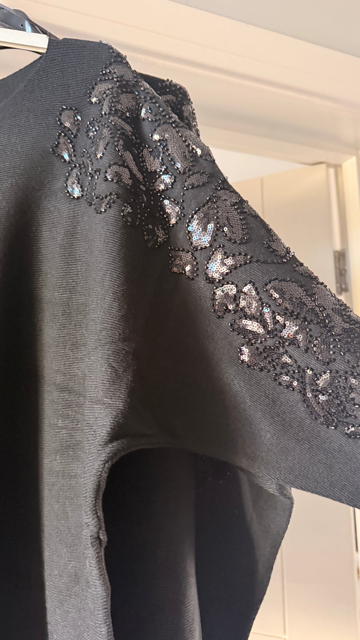 A close-up of a black knit garment hanging on a hanger, featuring an ornate floral pattern of sequins and beads across the shoulder and upper sleeve.