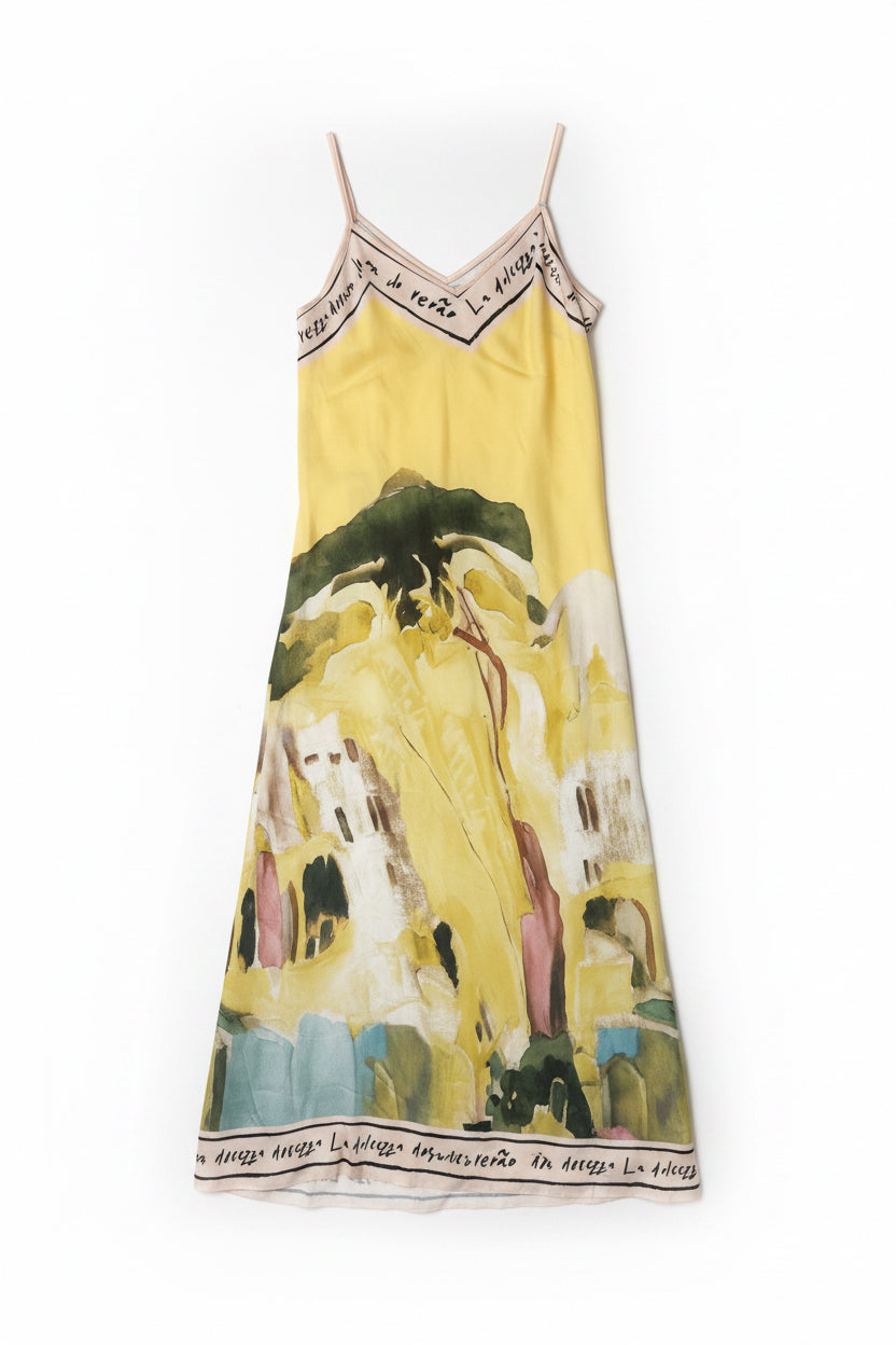 A yellow sleeveless maxi dress with a scenic landscape print, featuring thin straps and black text details on the neckline and hem, hanging on a white door.