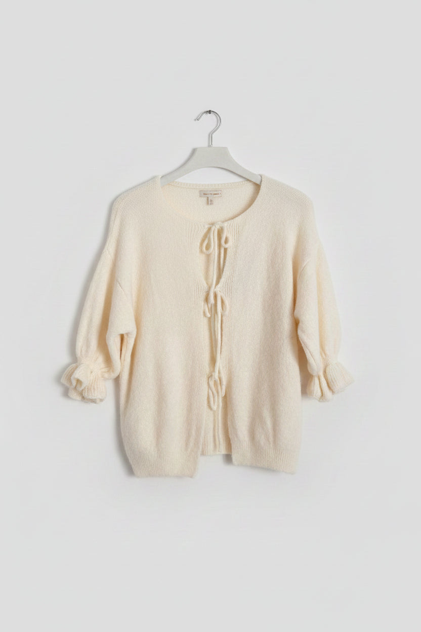 Cream-colored fuzzy knit cardigan sweater with tie-front closures and ruffled cuffs, hanging on a black hanger against a white paneled wall.