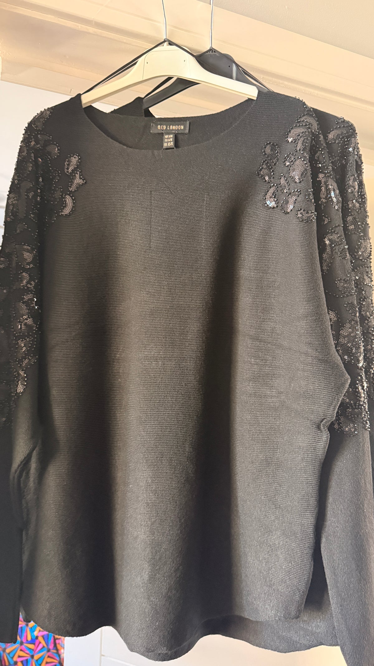 A black knit sweater with black lace applique on the shoulders and sleeves, hanging on a white hanger against a light-colored wooden door.