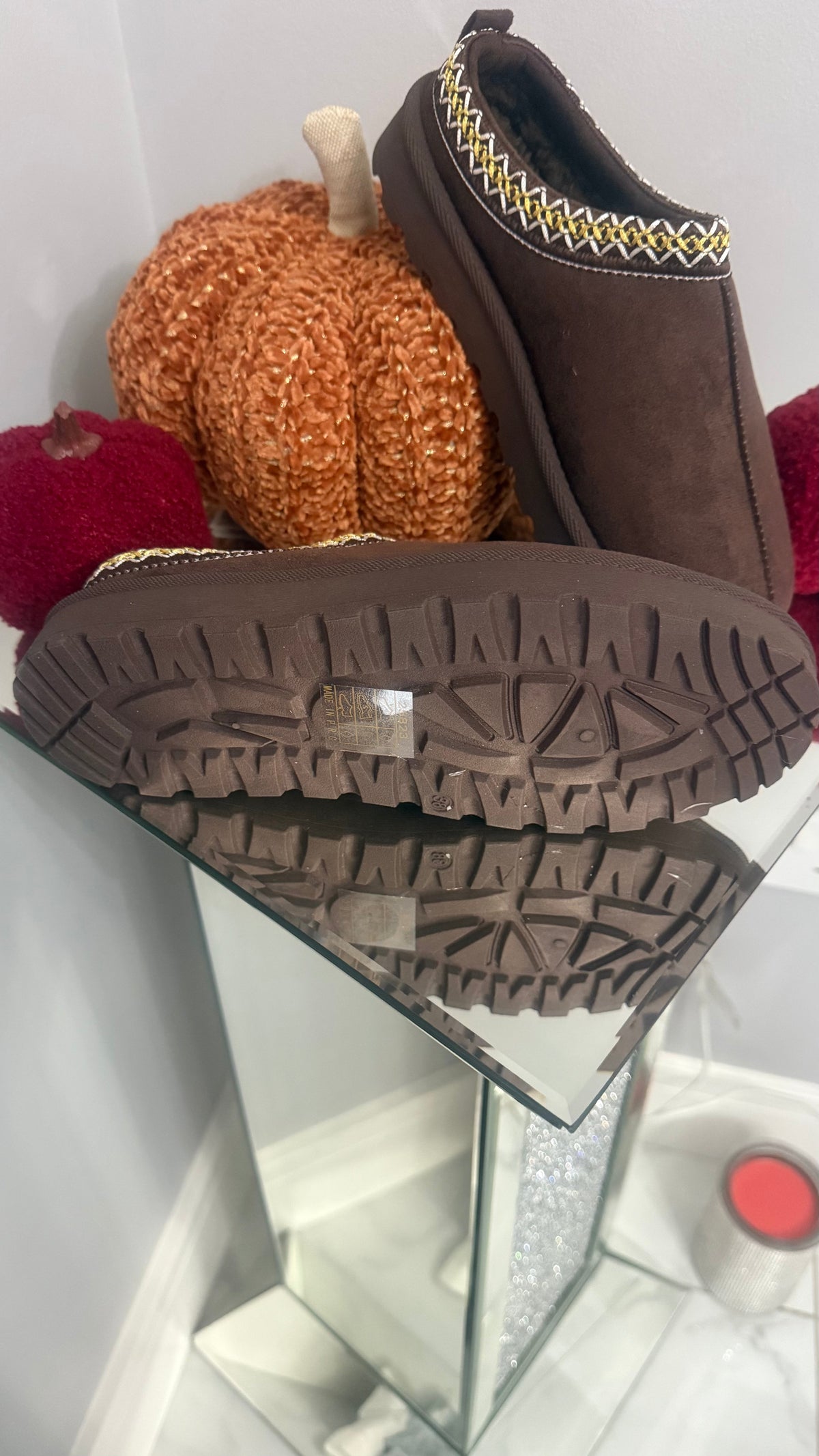 Brown slip-on shoes with a textured sole on a reflective surface, surrounded by decorative pumpkins.