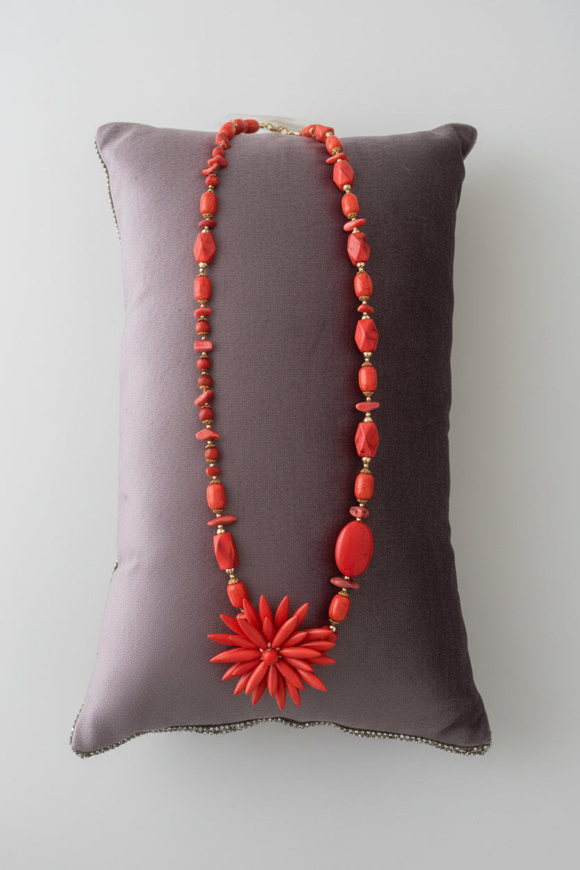 A long coral-red beaded necklace with a large flower-shaped pendant, displayed on a dark gray velvet pillow over a light gray marble surface.