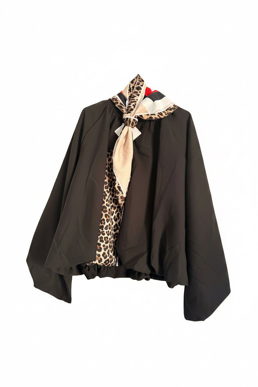 A black cape-style jacket with a beige and brown leopard-print collar and a beige neck scarf, hanging on a metal hanger against a white paneled wall.