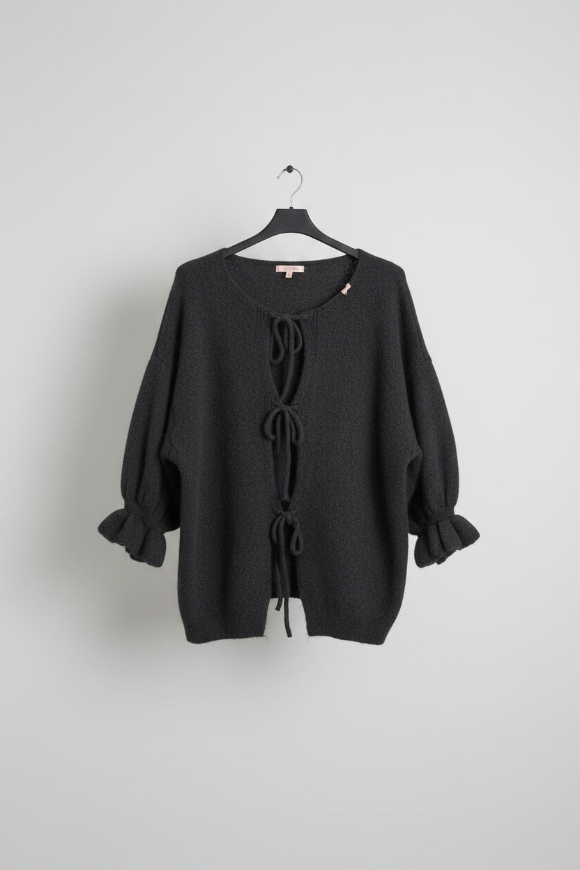 A dark gray fuzzy knit sweater with a round neckline, front tie closures, and ruffled elbow-length sleeves, hanging on a black hanger against a cream-colored tiled wall.