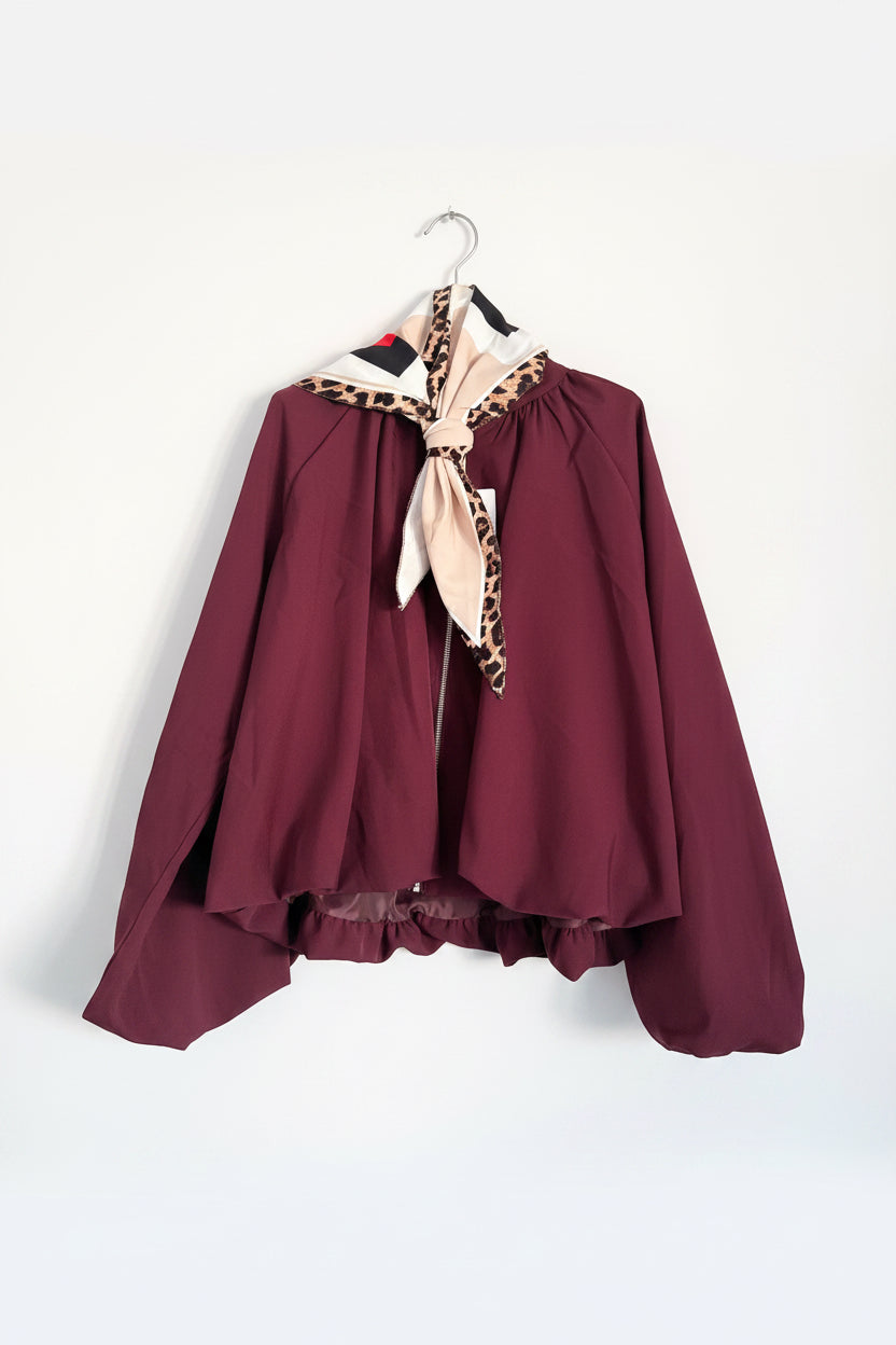A burgundy hooded cape or poncho-style garment hanging on a hanger against a white tiled wall, featuring a beige and black leopard-print trim along the hood and a matching beige neck tie.