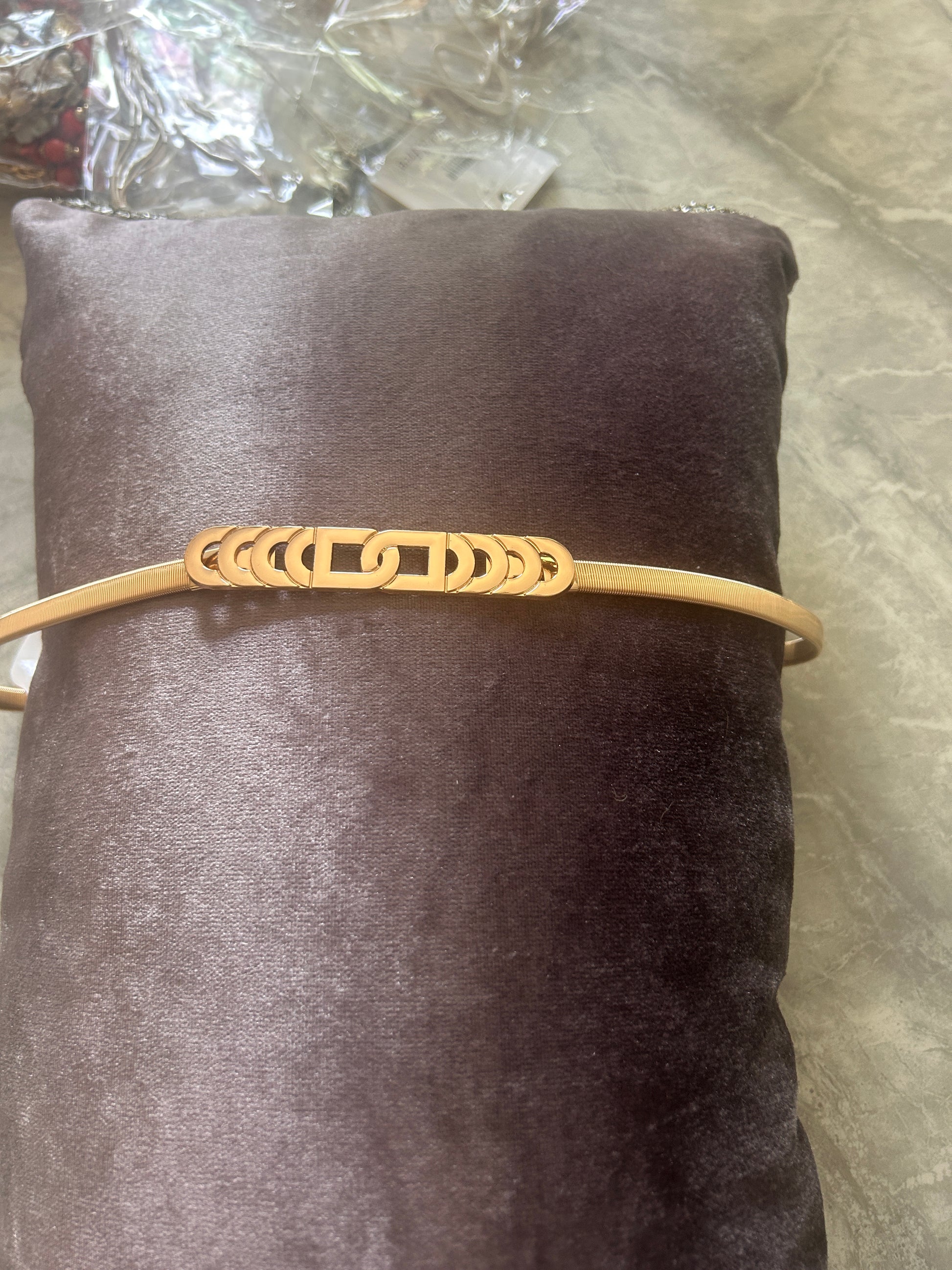 A gold-colored metal belt with a central cutout pattern of interlocking rings, displayed on a dark brown velvet cylindrical stand.