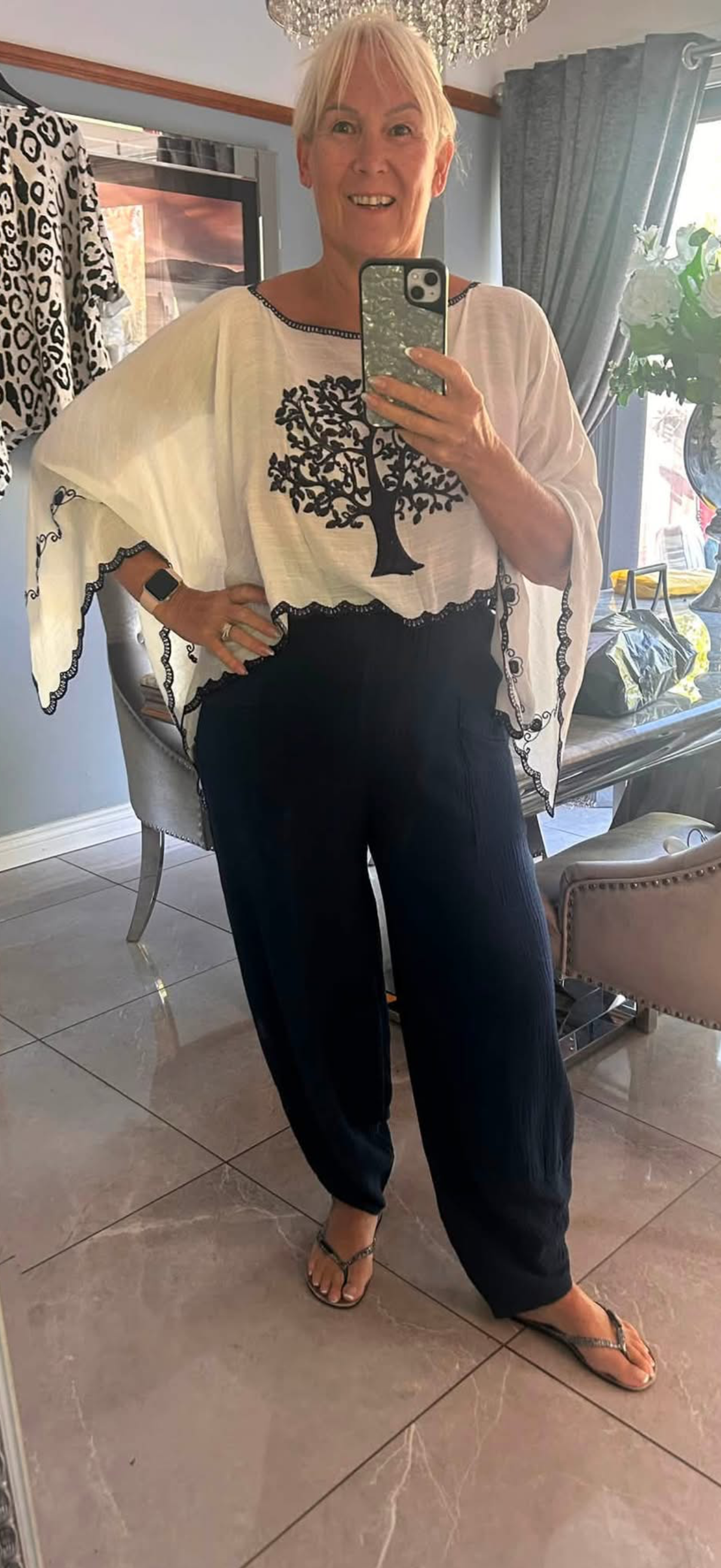 A woman taking a mirror selfie in a home setting, wearing a white top with a black tree graphic and black wide-leg pants.